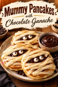 Stack of pancakes decorated with white chocolate drizzle to look like mummies, topped with chocolate ganache and candy eyes.